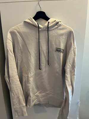 Beige hoodie från Hugo med tryck - Snygg beige hoodie från Hugo med svart logotyp på bröstet och ett stort, detaljerat tryck av en klassisk byggnad på ryggen. Hoodien har huva med svarta snören och är tillverkad i mjukt material. Perfekt för dig som gillar streetstyle och unika motiv.