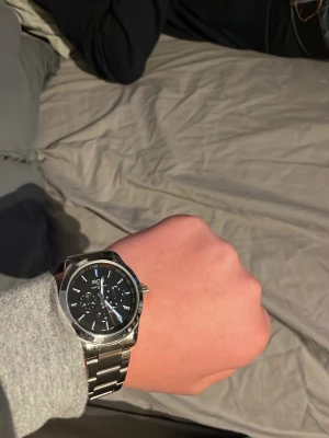Regal silverfärgad klocka - Stilren klocka från Regal med svart urtavla och silverfärgat armband i metall. Urtavlan har flera små visare och tachymeter runt kanten. Perfekt för dig som gillar klassiska accessoarer med modern touch.