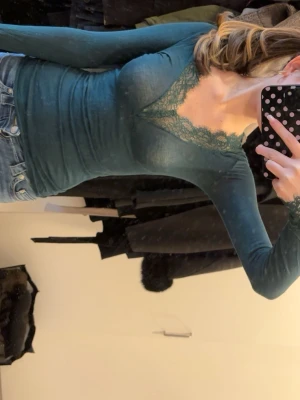 Intimissimi tröja - Snygg mörkgrön långärmad topp med v-ringning och dekorativ spets runt halsen. Toppens passform är tight och den är tillverkad i ett mjukt material som sitter bekvämt. Perfekt att matcha med jeans eller kjol.