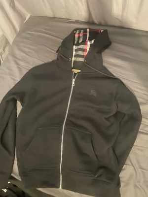 Svart Burberry hoodie med dragkedja - Svart hoodie från Burberry med hel dragkedja och klassiskt rutigt foder i huvan och insidan. Hoodien har två fickor framtill och en diskret Burberry-logga på bröstet. Perfekt för dig som gillar stilrena plagg med exklusiva detaljer.