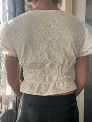 Vit broderad blus med v-ringning - Söt vit blus med broderade blommor och blad, smockad midja och v-ringning. Blusen har korta ärmar och är gjord i ett lätt, luftigt bomullstyg med hålbroderier. Perfekt som en sommartröja.