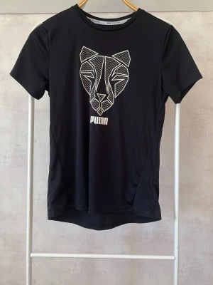 Svart Puma tränings t-shirt med grafiskt tryck - Svart t-shirt från Puma med ett stort, vitt grafiskt tryck av ett kattdjur på bröstet och Puma-logga under. T-shirten har rund halsringning och korta ärmar. Tillverkad i ett mjukt och stretchigt material som passar perfekt till träning.
