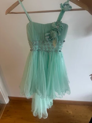 Mintgrön klänning med pärlor och blommor - Säljer en mintgrön klänning med tunna axelband, dekorerad med pärlor och blommor på överdelen. Klänningen har ett luftigt, transparent tyg och asymmetrisk kjol som ger ett drömmigt intryck. Perfekt för dig som vill sticka ut med en unik look. Passar 6/7 år.
