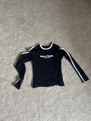 Svart långärmad topp med New York-tryck - Cool svart långärmad topp med vit text 'New York MANH NYC' på bröstet och siffran 97 på ryggen. Vit kant vid halsen och vita ränder längs ärmarna ger en sportig vibe. Perfekt för dig som gillar streetstyle och enkel design.