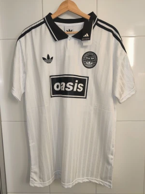 Oasis turnétröja / matchtröja 2025 - Säljer en turnétröja / matchtröja / fotbollströja för Oasis turné 2025. Vit tröja med svarta detaljer. Tröjan är en reprint helt ny och oanvänd. Storlek L.