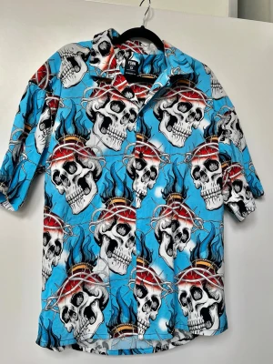 Cool skjorta  - En blå skjorta med dödskalletryck från märket FSBN (Fishbone), som säljs hos klädkedjan New Yorker. Skjortan är i klassisk hawaiimodell med kort ärm och knäppning framtill. Perfekt för dig som vill sticka ut med en cool och avslappnad stil – lika snygg till semester som till vardags! Strl M