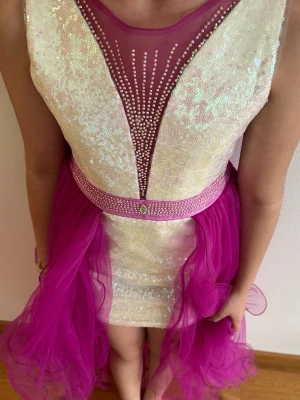 Glittrig rosa klänning med tyll - En kort klänning med ljus skimrande paljetter framtill, djup rosa mesh och dekorativa strassdetaljer. Midjan pryds av ett rosa band med strass och kjolen har ett lager av rosa tyll som ger volym och en dramatisk look. Dragkedja i ryggen och ärmlös modell. Passar 8/9 år.