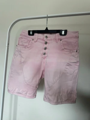 Ljusrosa shorts med knappar - Snygga ljusrosa shorts med fem knappar framtill och klassiska fickor både fram och bak. Shortsens material är mjukt och bekvämt, perfekt för varma dagar. De har en midwaist passform och en enkel, stilren design som är lätt att matcha.