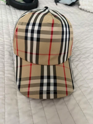 Burberry cap - Säljer en klassisk beige keps från Burberry med det ikoniska rutiga mönstret i svart, vitt och rött. Kepsen har böjd skärm och justerbar passform bak. Perfekt accessoar för dig som gillar stilrena och tidlösa detaljer.