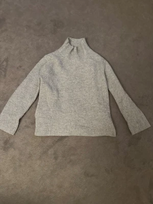 Ljusgrå stickad polotröja Little Goat - Mysig ljusgrå stickad polotröja från Soft Goat. Tröjan har ribbstickad struktur, hög krage och långa ärmar. Perfekt för kalla dagar och enkel att matcha med jeans eller kjol. Tröjan har en liten vit fläck på ena ärmen men det märks knappt.🤍