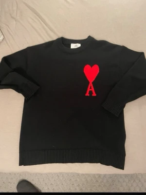 Svart stickad tröja med rött hjärta - Cool svart stickad tröja från AMI Paris med ett stort rött hjärta och bokstaven A på bröstet. Tröjan har rund hals, ribbade muddar och är tillverkad i mjukt material som passar perfekt för lager-på-lager. Perfekt för dig som gillar stilrena detaljer.