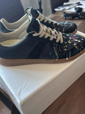 Maison Margiela skor -  Maison Margiela skor i svart läder och mocka med färgstänk i rött, blått, gult och vitt. Skorna har vita snören och beige innersula med logotyp. Perfekta nu inför sommaren! Självklart är dem äkta.