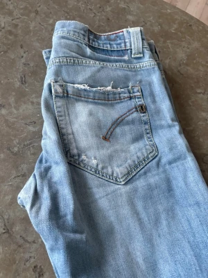 Dondup Jeans - Säljer ett par ljusblå jeans i klassisk femficksmodell med slitningar och råa detaljer på bakfickan. Jeansen har normal midja och raka ben. Perfekta för en avslappnad och trendig look. De är i storlek 34 men sitter som 30.