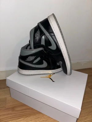 Air Jordan 1 Mid shadow gray - Säljer ett par Air Jordan 1 Mid shadow gray sneakers i svart och grått läder med klassisk Jordan-logga på sidan och tungan. Skorna har vita sulor och svarta skosnören. Perfekt för dig som gillar streetstyle och ikoniska sneakers.