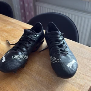 Svarta Puma Future fotbollsskor - Säljer ett par svarta Puma Future fotbollsskor med vita detaljer och logotyp. Skorna har snörning och mönstrad ovansida i syntetmaterial. Utrustade med fasta dobbar för bra grepp på planen. Perfekta för dig som vill ha stil och funktion på fotbollsplanen.