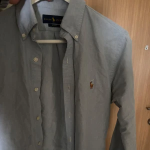 Blå skjorta från Ralph Lauren - Säljer en klassisk blå skjorta från Ralph Lauren i slim fit-modell. Skjortan har knappar framtill, button-down krage och den ikoniska broderade loggan på bröstet. Perfekt för en stilren look. Storlek S och mycket bra skick, inga defekter eller slitningar