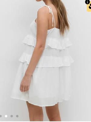 Vit volangklänning med knytband - Säljer en söt vit kortklänning med volanger och knytband på axlarna. Klänningen är ärmlös och har en luftig passform som är perfekt för varma dagar. Materialet ser ut att vara bomull eller linne, vilket gör den skön och lätt att bära. 🩷