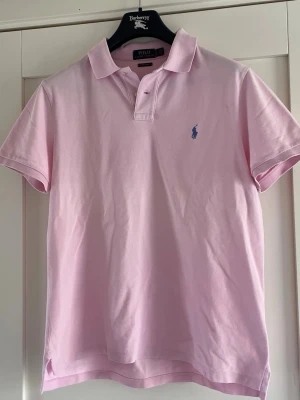 Ljusrosa pikétröja från Ralph Lauren - Säljer en ljusrosa pikétröja från Ralph Lauren med klassisk krage och två knappar framtill. Tröjan har den ikoniska blå loggan broderad på bröstet och är i en slim fit-modell. Tillverkad i mjuk bomull som känns skön mot huden.