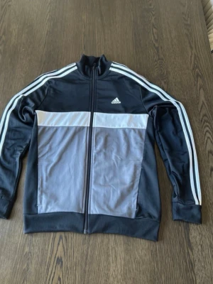 Svart och grå Adidas träningsjacka - Snygg träningsjacka från Adidas i svart och grått med vita ränder längs ärmarna och dragkedja framtill. Jackan har ståkrage, två sidofickor och Adidas-logga på bröstet. Tillverkad i ett mjukt och stretchigt material som passar perfekt till träning eller chill.
