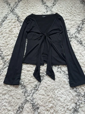 Svart långärmad topp från Gina Tricot - Svart långärmad topp från Gina Tricot med snygg knytning framtill och v-ringad hals. Toppens material är mjukt och stretchigt, perfekt för att skapa en cool och trendig look. Passformen är normal och ärmarna är lite utsvängda.