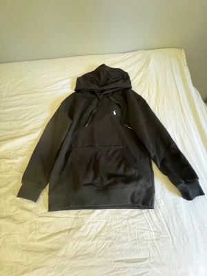 Svart hoodie från Polo Ralph Lauren - Säljer en svart hoodie från Polo Ralph Lauren med klassisk logga broderad på bröstet. Hoodien har huva med dragsko, stor magficka och ribbade muddar. Perfekt för dig som gillar stilren design och ikoniska märken.