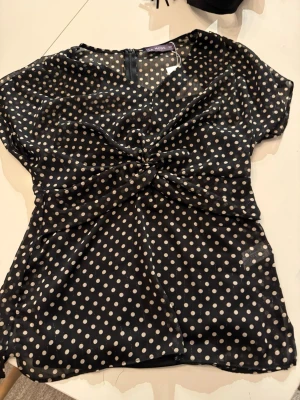Svart prickig blus från Mode - Snygg svart blus från Mode med vita prickar och twistad detalj framtill. Blusen är gjord i ett tunt, transparent material och har korta ärmar. Perfekt för dig som gillar retrostil och vill ha något lätt och luftigt.