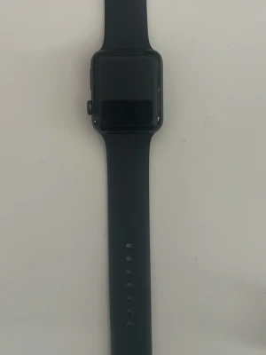 Svart Apple Watch med silikonband - Säljer en svart Apple Watch series 3 40 mm med ett stilrent silikonband. Klockan har en rektangulär touchskärm och en rund knapp på sidan. Perfekt för att hålla koll på notiser, träning och hälsa. Bandet är justerbart och passar de flesta handleder.
