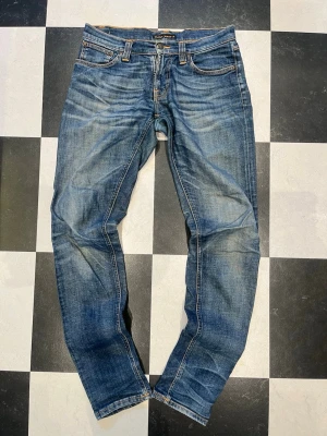 Blå jeans från Nudie Jeans - Säljer ett par klassiska blå jeans från Nudie Jeans med snygga detaljer och slim passform. Materialet är bomull och de har en cool faded look som ger en avslappnad vibe.