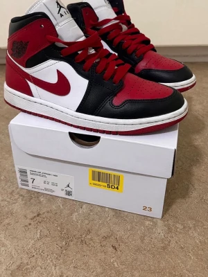 Air Jordan 1 Mid röd/svart/vit - Säljer ett par Air Jordan 1 Mid sneakers i klassisk röd, svart och vit färgkombination. Skorna har läderöverdel, röda skosnören och ikoniska Swoosh-loggan på sidan. Perfekta för dig som gillar streetwear och vill sticka ut med en tidlös modell.