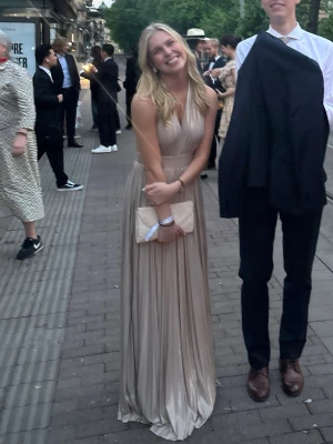 Guldig långklänning med halterneck - Elegant guldig långklänning i glansigt material med halterneck och öppen rygg. Klänningen har knyt i nacken och ett vackert fall med plisserad kjol. Perfekt för tex bal eller andra fina event.🥰Köpt för ca 1000 sek 