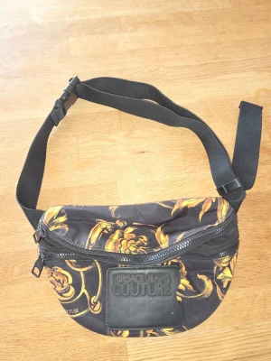 Axelväska svart/guld - Snygg axelväska i svart tyg med ikoniskt barockmönster i guld. Väskan har justerbart band, två dragkedjefack och en stor logopatch framtill. Perfekt för dig som vill ha en trendig och praktisk accessoar.