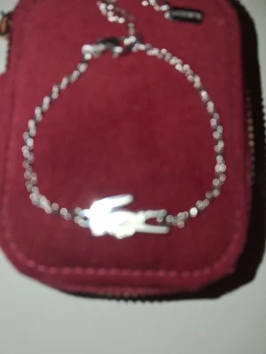 Silverfärgat armband från Lacoste - Snyggt armband i silverfärg från Lacoste med tunn kedja och en liten krokodilberlock. Armbandet har ett diskret Lacoste-märke vid låset och passar perfekt för dig som gillar stilrena accessoarer. Materialet är metall och designen är enkel men ändå trendig.