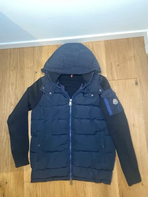 Mörkblå cardiganjacka från Moncler - Snygg mörkblå cardiganjacka från Moncler med vadderad front och stickade ärmar. Jackan har huva, dragkedja framtill och en ikonisk Moncler-logga på ärmen. Materialmix med dun i framstycket och mjukt tyg på ärmar och rygg. Perfekt för lager på lager och streetstyle.