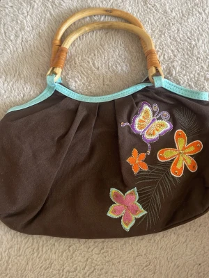 Brun tygväska med bambuhandtag, 2000-stil - Söt brun handväska i tyg med ljusblå kant och broderade blommor och fjäril i färgglada nyanser. Väskan har runda handtag i bambu och en mjuk, rundad form. Perfekt för dig som gillar boho-stil och unika detaljer.