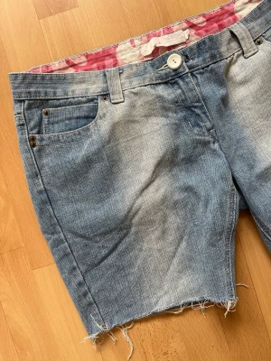 Vintage Y2K jeansshorts - Coola ljusblå denimshorts, från börjar ett par långa jeans. Har en sliten look med råa, fransiga kanter nertill. Fina broderade detaljer på bakfickorna, en vit knapp fram och ett supergulligt rosa kamouflagemönstrat foder på insidan av linningen (syns om du viker ner kanten)✨