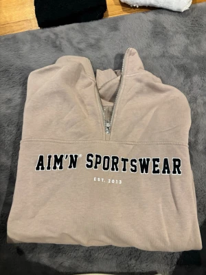 Beige Aim'n Sportswear hoodie - Säljer en beige hoodie från Aim'n Sportswear med halv dragkedja och svartvit text framtill. Hoodien har ribbade muddar och är tillverkad i mjukt sweatshirtmaterial. Perfekt för en avslappnad och sportig look.