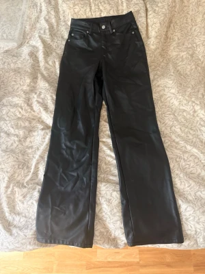 Svarta vida jeans i skinnimitation - Säljer ett par svarta jeans med hög midja och vida ben i skinnimitation. Klassisk femficksmodell med knapp och dragkedja framtill. Perfekta för dig som vill ha en edgy och trendig look.