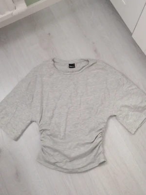 Ljusgrå topp från Gina Tricot - Säljer en ljusgrå topp från Gina Tricot med korta ärmar och rund hals. Toppen har en snygg rynkning i sidan som ger en cool twist till den annars enkla looken. Perfekt att styla med jeans eller kjol för en trendig vibe.