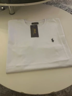 Vit t-shirt från Polo Ralph Lauren - Snygg vit t-shirt från Polo Ralph Lauren med klassisk rund hals och den ikoniska loggan broderad på bröstet. Tillverkad i mjukt bomullstyg som känns skönt mot huden. Perfekt basplagg för en clean och stilren look.