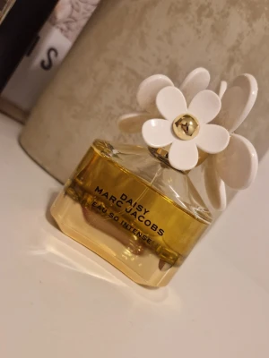 Daisy Eau So Intense parfym - Säljer en Daisy Eau So Intense parfym från Marc Jacobs. Flaskan är guldfärgad med en unik design och har vita blommor på korken. Perfekt för dig som gillar söta och fräscha dofter och vill ha en snygg parfymflaska på hyllan.