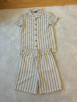 Randig pyjamas med kort ärm och shorts - Snygg pyjamas med kortärmad skjorta och shorts i vitt med svarta och beige ränder. Skjortan har knappar framtill och krage, shortsen har resår och dragsko i midjan. Perfekt för varma nätter och chill hemma.