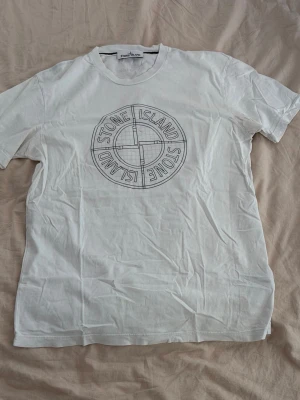 Vit Stone Island t-shirt med tryck - Vit t-shirt från Stone Island med stort rundat logotryck på bröstet. Klassisk passform med korta ärmar och rund halsringning. Tillverkad i mjuk bomull, perfekt för en clean och stilren look.