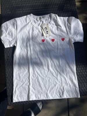 Vit t-shirt med röda hjärtan PLAY - Vit t-shirt från PLAY med tre små röda hjärtan broderade på bröstet. Klassisk rund hals och korta ärmar. Perfekt för dig som gillar enkel men söt design. Tillverkad i mjuk bomull för skön känsla hela dagen.