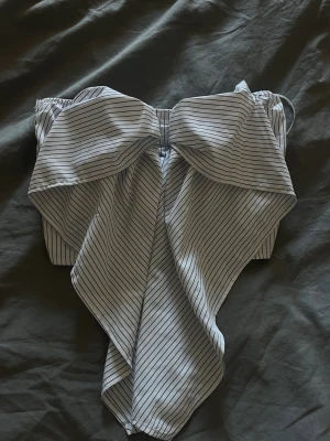 Randig bandeau topp med rosett - Säljer en snygg bandeau topp i vitt med tunna svarta ränder och stor rosettdetalj framtill. Toppen är axelbandslös och har smockad rygg för skön passform. Perfekt för dig som vill sticka ut med en unik och trendig look.