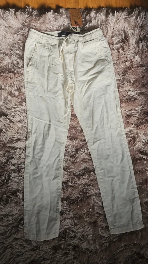 Vita linnebyxor med dragsko - Snygga vita linnebyxor med resår och dragsko i midjan. Byxorna har raka ben, sidofickor fram och två fickor bak. Perfekta för en avslappnad och stilren look. Materialet är lätt och luftigt, vilket gör dem sköna att bära under varmare dagar. 