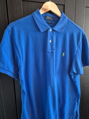 olo Ralph Lauren piké  - Klassisk blå pikétröja från Polo Ralph Lauren med krage, korta ärmar och knappar framtill. Tröjan har den ikoniska gröna logotypen broderad på bröstet och är tillverkad i bomull. Perfekt för en stilren och avslappnad look.
