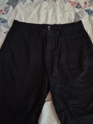 Hm linne kostymbyxor  - Snygga svarta chinos från H&M med klassisk rak passform. Byxorna har sidofickor fram, två bakfickor med knapp och hällor för bälte. Materialet ser ut att vara bomullsmix som ger en mjuk och bekväm känsla. Perfekta för en clean och stilren look.