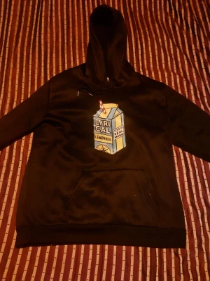 Svart hoodie med Lyrical Lemonade tryck - Säljer en svart hoodie med en stor färgglad Lyrical Lemonade-mjölkpaketgrafik på bröstet. Hoodien har huva, magficka och ribbade muddar. Perfekt för dig som gillar streetwear och vill sticka ut med en unik design.