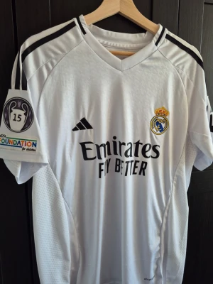 Real Madrid Bellingham matchtröja - Säljer en vit Real Madrid matchtröja från Adidas med svarta detaljer och tryck. Tröjan har Bellingham och nummer 5 på ryggen, samt klubbmärke och sponsorlogga framtill. UEFA Foundation-märke på ärmen. Perfekt för fotbollsfans!