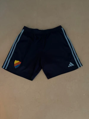 Blå träningsshorts från Adidas M - Mörkblå träningsshorts från Adidas med ljusblå ränder längs sidorna och klubbmärke i regnbågsfärger på ena benet. Shortsen har elastisk midja och är tillverkade i ett lätt, snabbtorkande material. Perfekta för fotboll eller gym. Shortsen är använda ett par gånger men fungerar felfritt. ABS! Pris går att diskutera! 